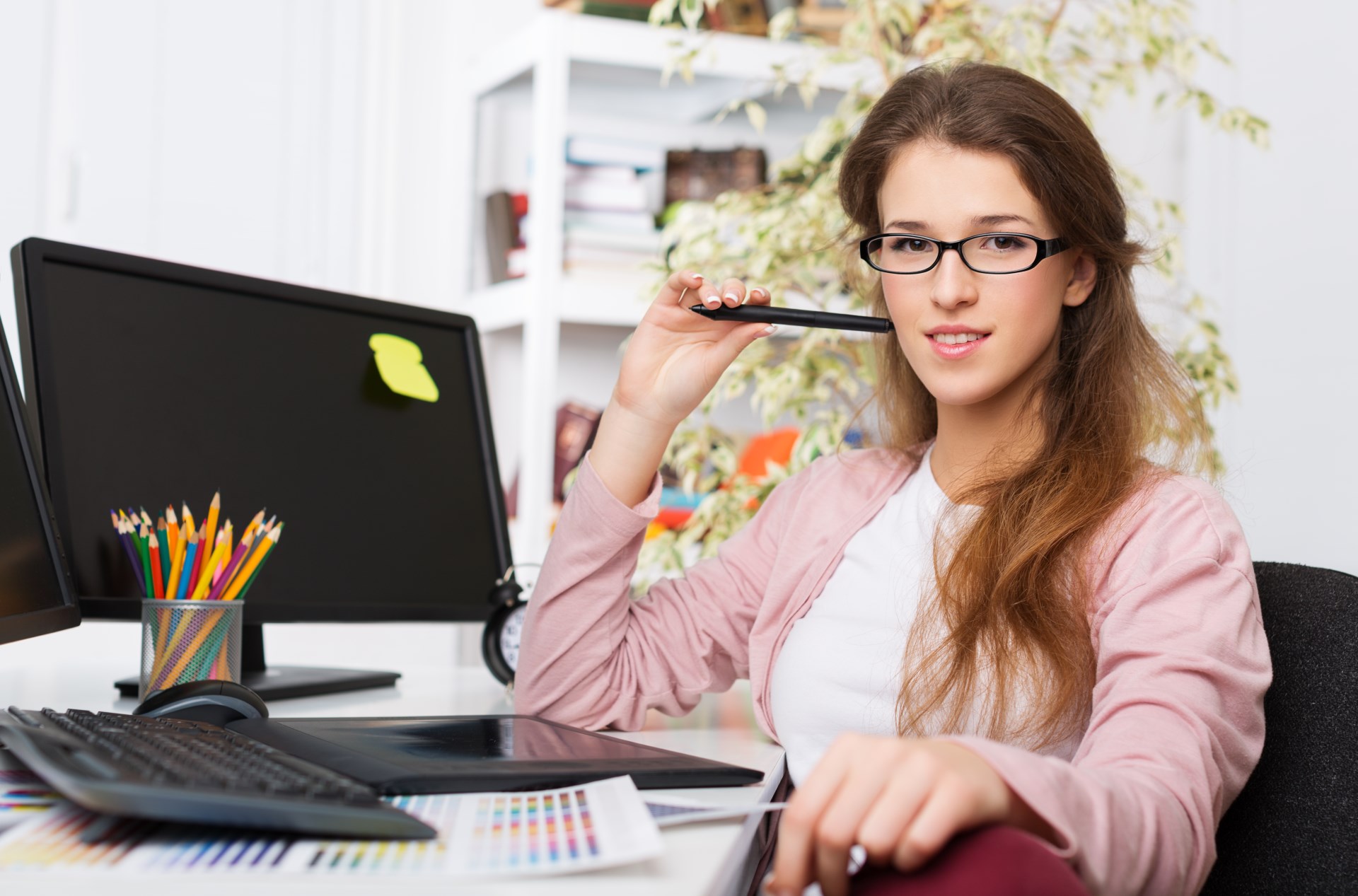 Frau mit Brille hält Stift, sitzt am Schreibtisch mit Computer, Tastatur und Buntstiften.